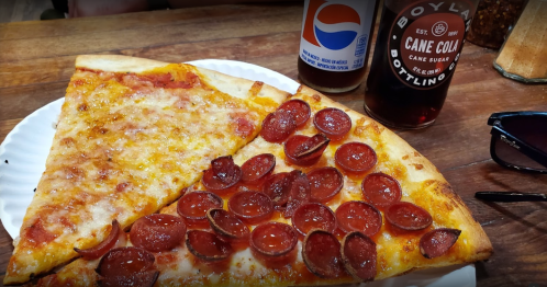 A slice of pepperoni pizza on a plate, accompanied by bottles of soda and sunglasses on a wooden table.