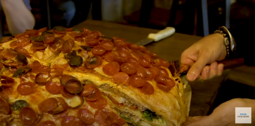 A large, layered pizza topped with pepperoni and mushrooms, being sliced by a person.