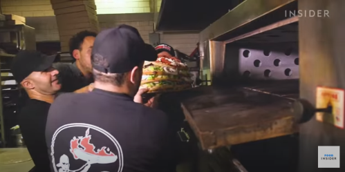 Four people are placing a large stack of pizzas into a commercial oven in a busy kitchen.