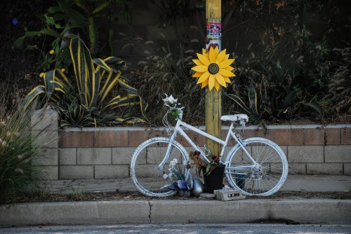 A white bicycle adorned with flowers and a sunflower decoration, leaning against a pole in a garden setting.