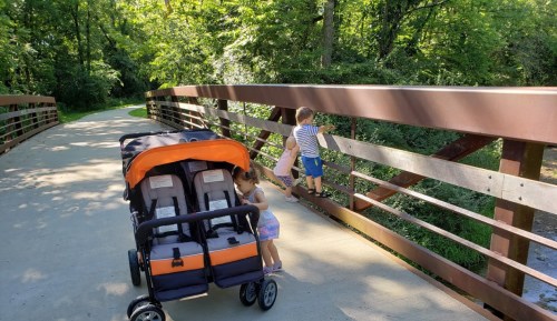 Two children play near a stroller on a bridge surrounded by greenery, while another child climbs on the railing.