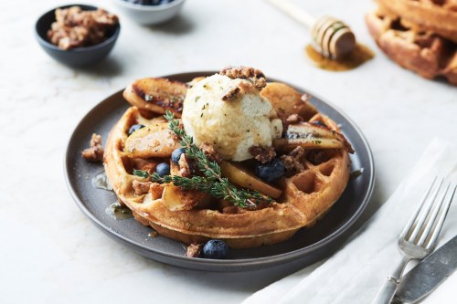 A plate of waffles topped with ice cream, sautéed apples, blueberries, and nuts, drizzled with honey.