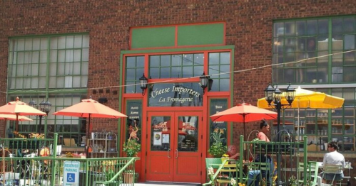 A charming cheese shop with outdoor seating, featuring orange umbrellas and a brick facade.