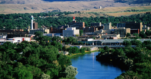 A scenic view of a city along a river, surrounded by greenery and hills in the background.