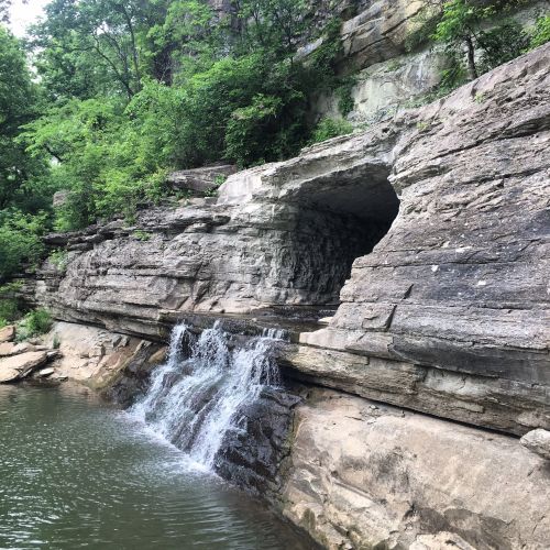 A rocky cliff with a small waterfall flowing into a calm body of water, surrounded by lush green trees.