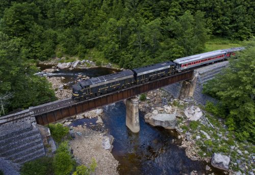 A train crosses a bridge over a river, surrounded by lush green trees and rocky terrain.