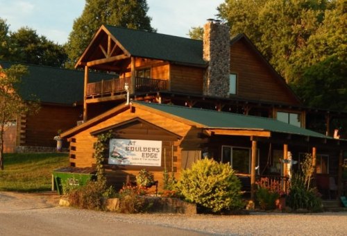 A rustic log cabin with a stone chimney, surrounded by greenery and a gravel driveway, featuring a sign that reads "At Builder's Edge."