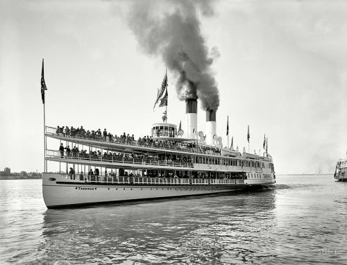 A historic steamboat with multiple smokestacks, filled with passengers, sailing on a river. Flags wave from the deck.