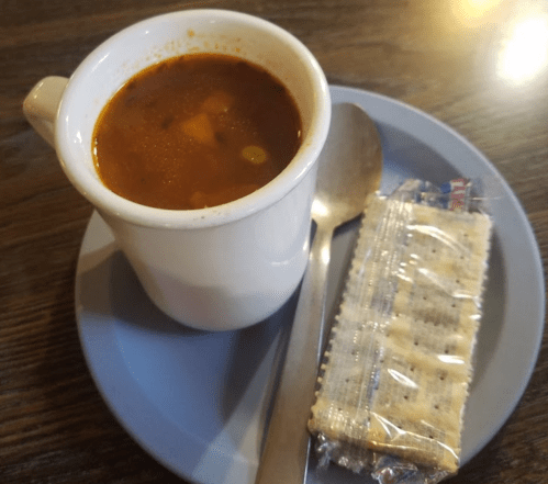 A cup of soup with vegetables, a spoon, and a pack of crackers on a blue plate.