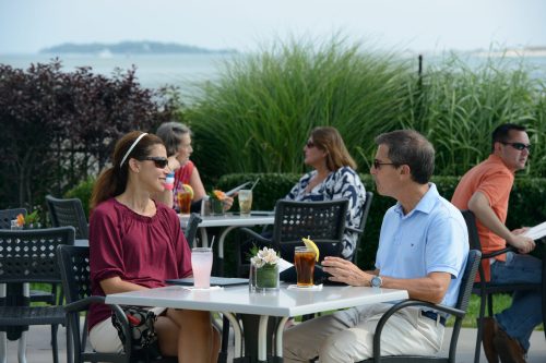 Two people sit at a table outdoors, enjoying drinks, with others in the background and a scenic view of water and greenery.