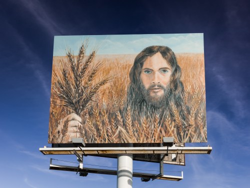 Billboard featuring a painted portrait of a man with long hair and a beard, holding wheat in a golden field.