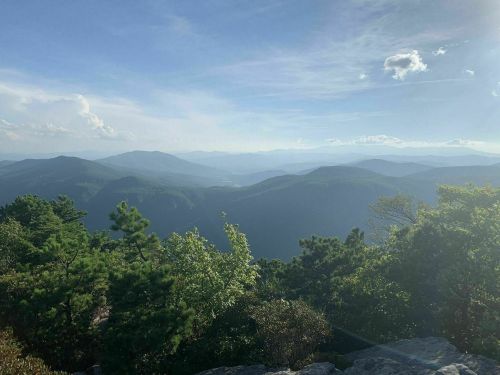 A scenic view of layered mountains under a blue sky, with greenery in the foreground and soft clouds above.