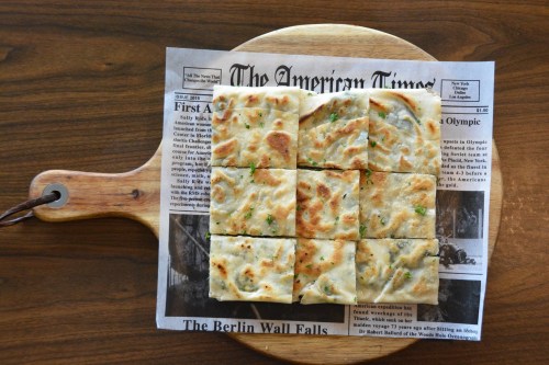 A wooden board with square pieces of flatbread arranged on a newspaper, showcasing a crispy, golden-brown texture.