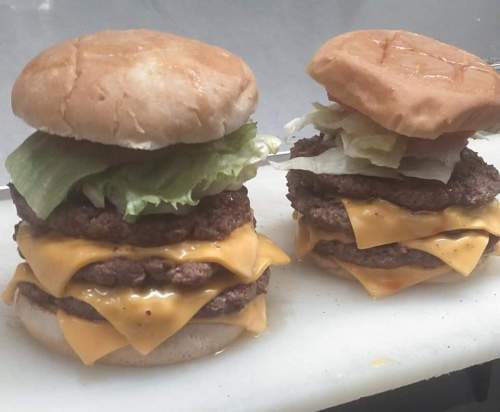 Two stacked burgers with three beef patties, melted cheddar cheese, lettuce, and tomato, served on toasted buns.