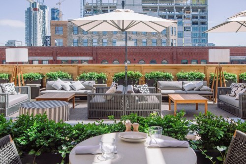 Rooftop terrace with modern seating, umbrellas, and greenery, overlooking a city skyline with construction in the background.