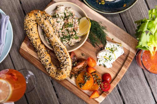 A wooden platter with a bagel, smoked fish spread, fresh herbs, cherry tomatoes, and a creamy dip, alongside drinks.