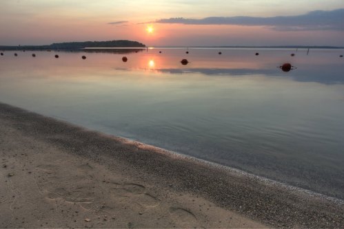 Sunset over calm water with a sandy shore and buoys floating in the distance, creating a serene atmosphere.