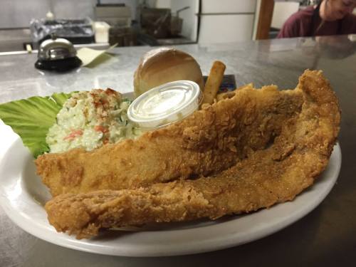 A plate featuring two pieces of fried fish, coleslaw, a roll, and a small container of sauce on a bed of lettuce.