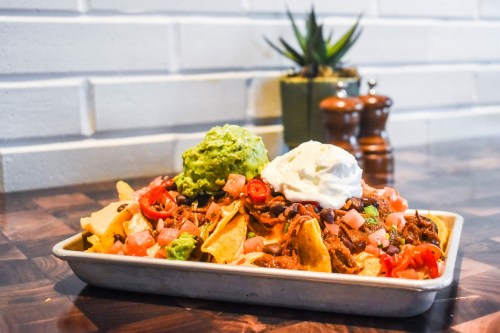 A platter of nachos topped with guacamole, sour cream, diced tomatoes, and jalapeños on a wooden table.