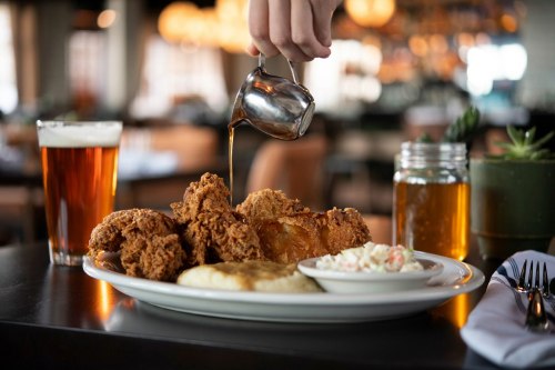 A hand pours sauce over a plate of fried chicken, served with coleslaw and mashed potatoes, alongside two glasses of beer.