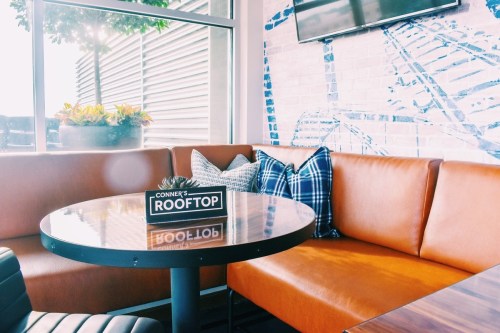 Cozy rooftop seating area with a round table, brown leather couch, and decorative pillows against a light brick wall.