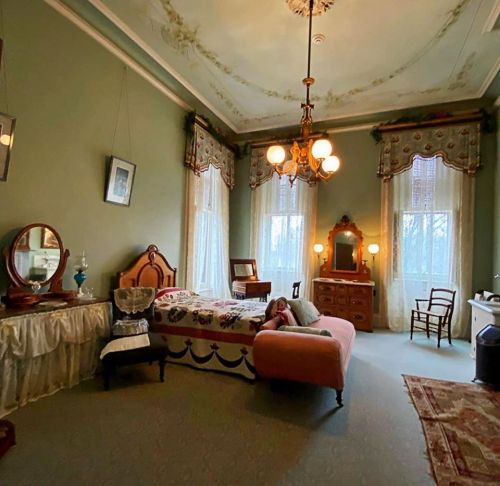 A vintage bedroom with a bed, dresser, mirror, and elegant decor, featuring large windows and soft lighting.
