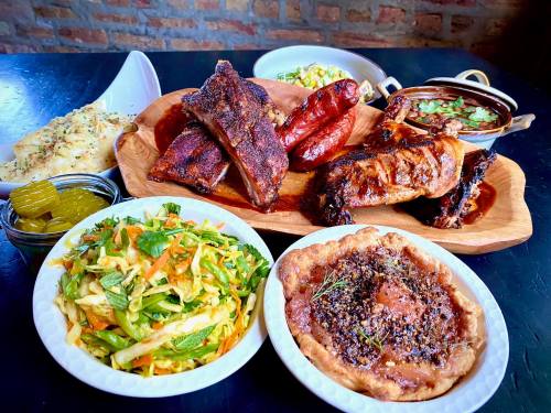 A wooden platter with assorted meats, including ribs and sausage, alongside sides of salad, pickles, and a savory pie.