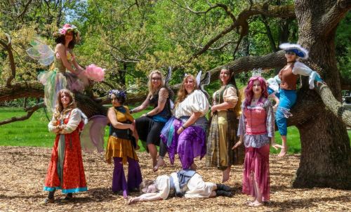 A group of people in colorful costumes poses playfully on and around a large tree in a sunny park setting.