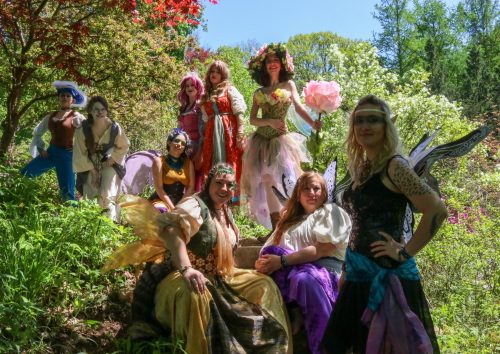 A group of women dressed as fairies and fantasy characters poses in a lush, colorful garden setting.