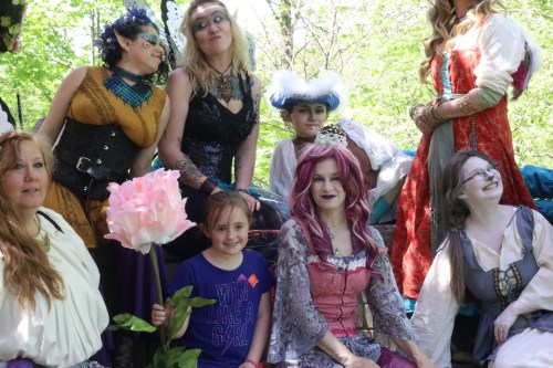 A group of people in colorful costumes poses together outdoors, with a child holding a large flower in the front.