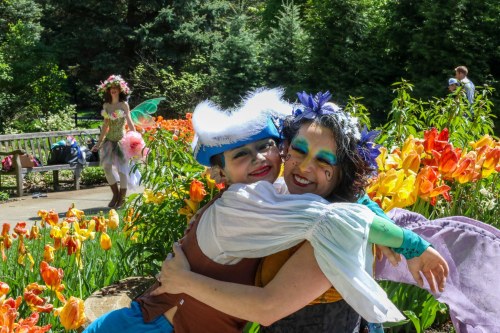 Two people in colorful costumes embrace in a flower garden, with a third person in the background among the blooms.