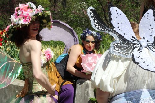 Three individuals in colorful, whimsical costumes with floral and butterfly themes, surrounded by greenery and flowers.
