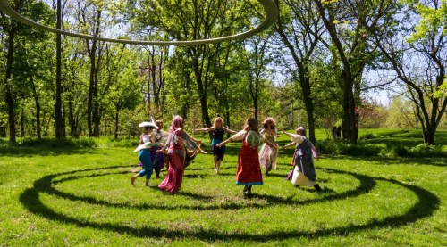 A group of people in colorful costumes dance in a circle on a grassy field surrounded by trees.
