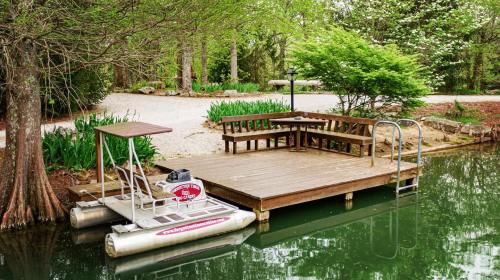 A wooden dock with a bench and table beside a calm, green pond surrounded by trees and blooming plants.