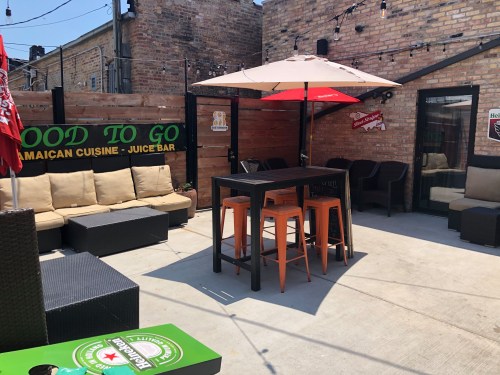 Outdoor seating area with a table and chairs, surrounded by brick walls and umbrellas, featuring Jamaican cuisine signage.