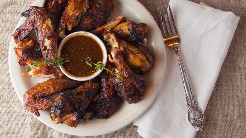 A plate of grilled chicken wings served with a small bowl of sauce and a fork on a linen tablecloth.