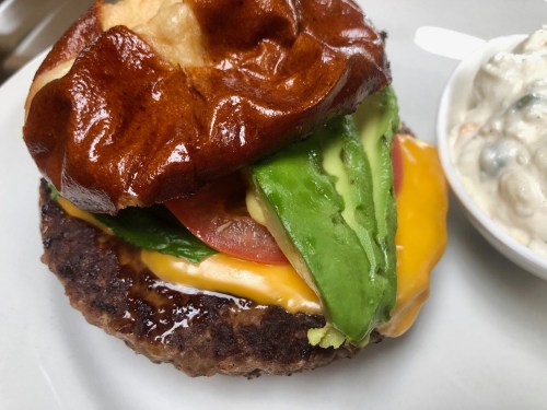 A close-up of a juicy burger with a pretzel bun, melted cheese, lettuce, tomato, and a side of creamy dip.