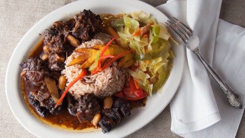 A plate of rice and peas topped with stewed beef, fried plantains, and a side of sautéed vegetables.