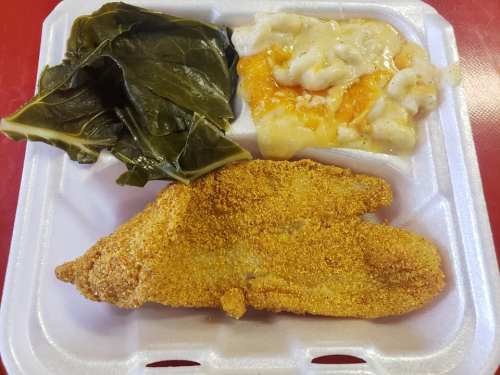 A styrofoam container with fried fish, collard greens, and macaroni and cheese.