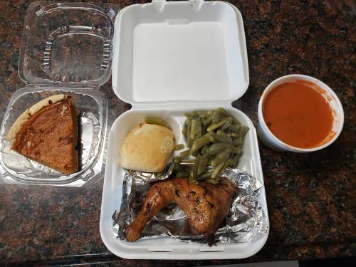 A takeout meal featuring a baked chicken leg, green beans, a roll, tomato soup, and a slice of pie in a white container.