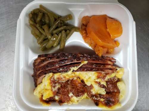 A styrofoam food container with sliced meatloaf, macaroni and cheese, green beans, and glazed carrots.