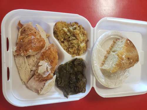 A takeout container with sliced chicken, stuffing, collard greens, and a piece of cornbread.