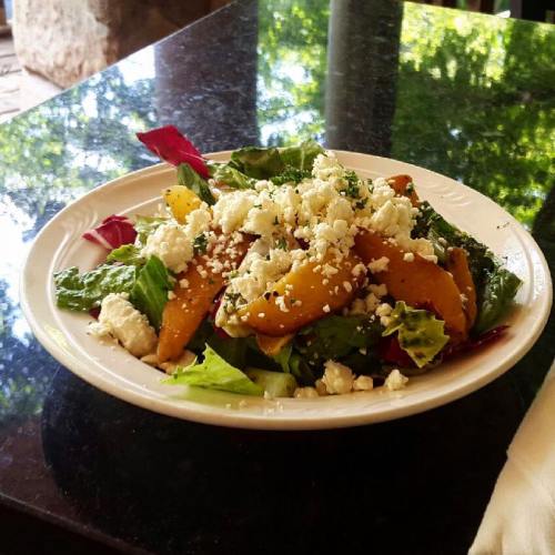 A fresh salad with mixed greens, roasted squash, and crumbled feta cheese on a white plate, set on a dark table.
