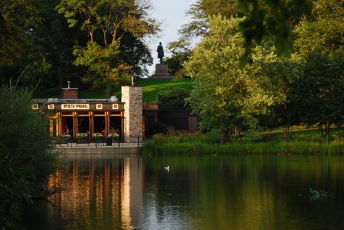 A serene park scene featuring a pond, a gazebo, and a statue on a hill surrounded by lush greenery.