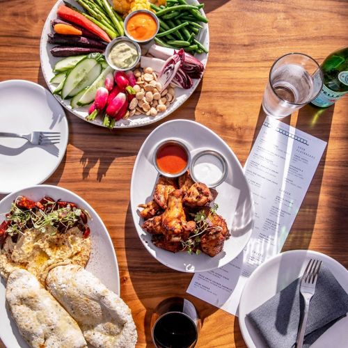 A table set with plates of colorful vegetables, chicken wings, and a menu, with drinks and utensils nearby.