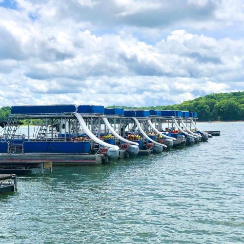 A row of boats with slides docked on a calm lake under a partly cloudy sky.