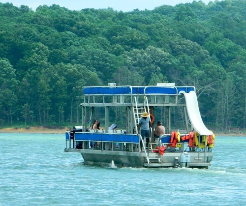 A party boat with people enjoying the water, featuring a slide, surrounded by green trees and calm water.