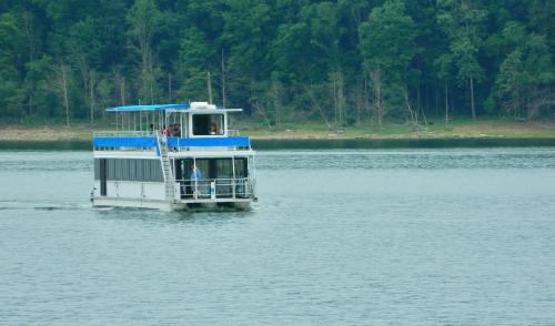 A boat with a blue roof glides across a calm lake, surrounded by lush green trees on the shore.