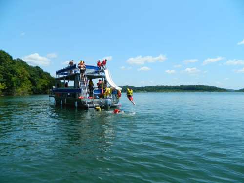 A group of people on a boat with a slide, enjoying a sunny day on the water, with one person jumping in.