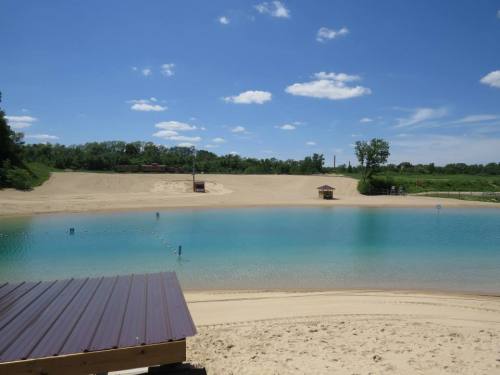 A serene lake with clear blue water, sandy shores, and a bright blue sky dotted with fluffy clouds.